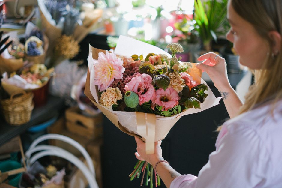 A woman with long, light brown hair, wearing a light pink shirt, is holding a bouquet of fresh flowers inside a flower shop. The bouquet features pink and peach roses, a large pale pink dahlia, small 