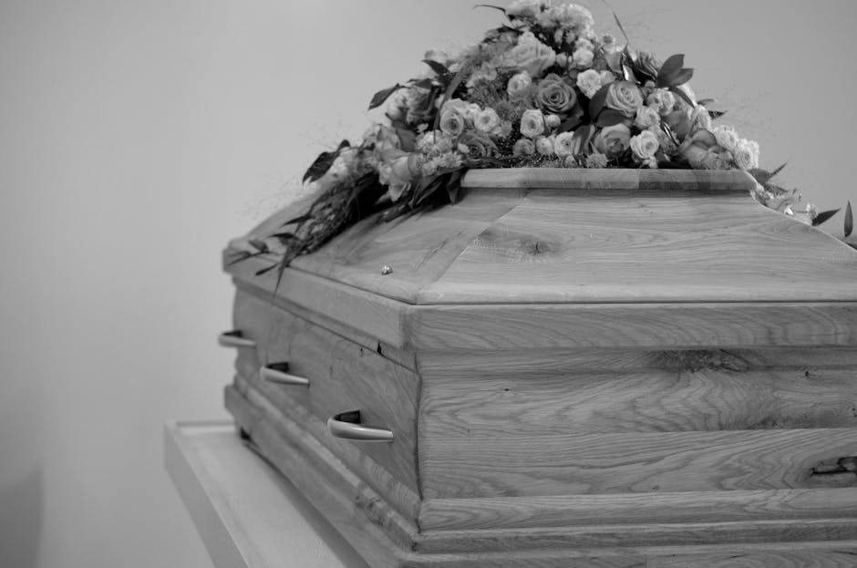 A wooden coffin resting on a white table, decorated with a floral arrangement featuring roses, carnations, and various greenery, arranged across the top and sides of the coffin. The flowers are in sof