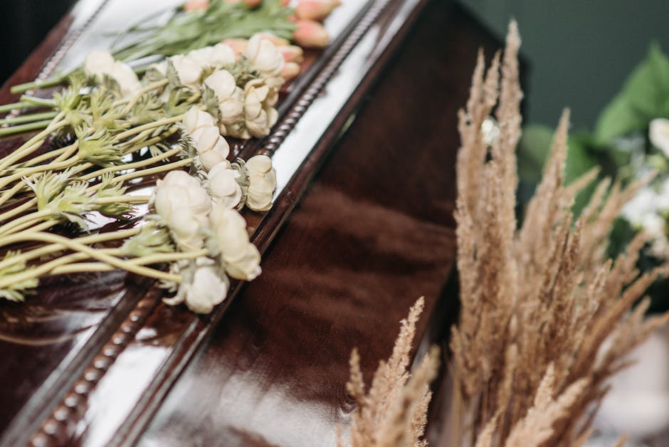 A close-up view of a floral arrangement featuring a bouquet of white ranunculus flowers and pink tulips, laid on a polished dark wooden surface. The bouquet is wrapped in green floral paper with a mat