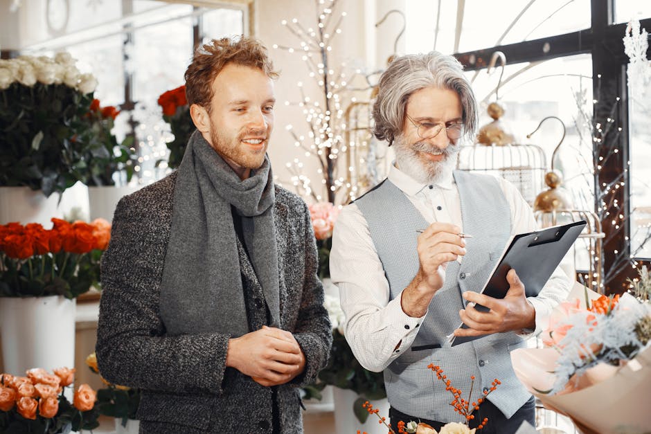 A flower delivery professional in a flower shop, wearing a light grey vest and white shirt, uses a black tablet to check an order while showing a bouquet of mixed flowers to a male customer. The bouqu