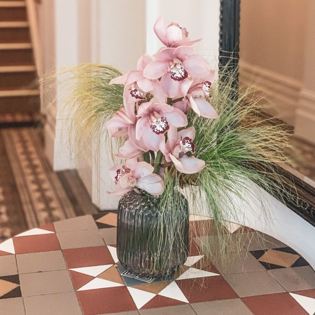A floral arrangement featuring a tall bouquet of pink and white orchids with intricate markings on the petals, placed in a dark ribbed glass vase. The orchids are complemented by wispy green ornamenta