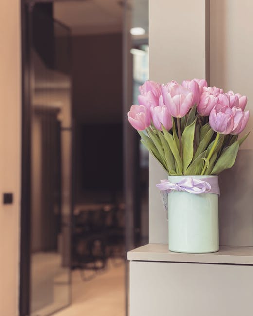 A vase of fresh pink tulips with green stems and leaves, arranged in a cylindrical light green ceramic container tied with a pale purple ribbon, placed on a white surface near a wall. The tulips are f