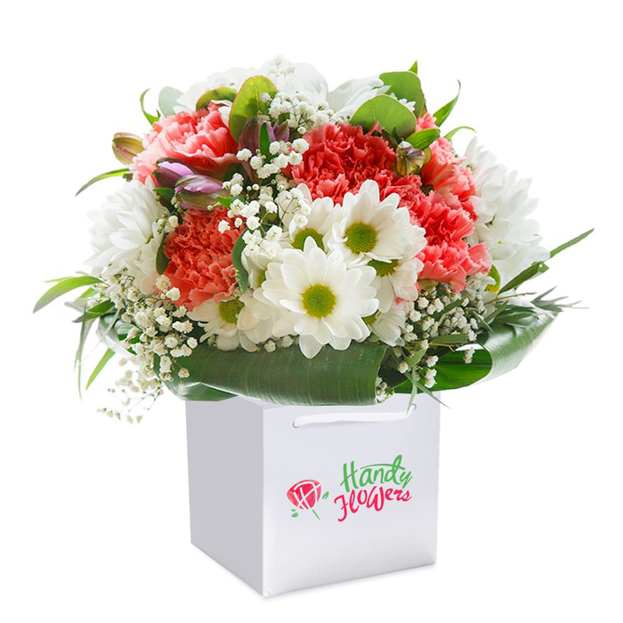 Beautifully upright in a crisp, modern white gift bag, this vividly arranged bouquet is a showcase of expert Cobham floristry, set for delivery across the village and its leafy surrounds. Soft, peach-coral carnations, their frilly petals imparting both volume and charm, are interspersed with clean white daisies crowned by bright green centers-a joyful pairing that recalls the cheerful flora of Cobham's countryside lanes. Throughout the bouquet, airy clusters of white gypsophila (baby's breath) offer gentle movement and softness, balancing the more robust forms. Tucked discreetly are a handful of closed purple-tipped buds, suggesting early spring along the riverbanks and the anticipation of new blooms. The whole arrangement is embraced by glossy, dark green foliage, the leaves arching gracefully and giving the piece a beautifully rounded form. A set of white ribbon handles on the gift bag makes for effortless presentation, whether for a Cobham High Street celebration, a thoughtful birthday surprise, or a special gesture for a neighbour. With a pristine white background setting the stage, the bouquet's abundant colour and exquisite detail shine through. Every element reflects a sense of freshness, proficiency, and heartfelt care that's synonymous with local Cobham florists.