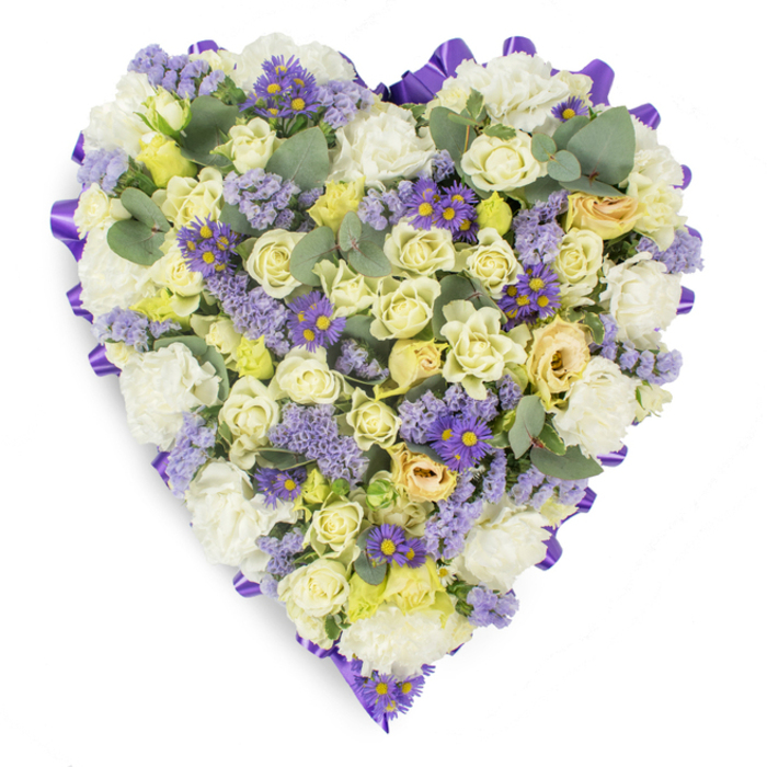A lovingly assembled heart-shaped tribute photographed on a clean white background, where every bloom and leaf is chosen to comfort and reassure. The densely arranged centre is dominated by cream and pale yellow roses, ranging from neat buds to gently unfurling flowers whose satiny surfaces catch a soft studio light; among them, ruffled white carnations add body and a traditional, consoling texture. Interspersed lavender statice breathes a faint, meadow-like lightness through the composition, while small deep-violet, aster-style flowers with bright yellow centres provide vivid, grounding accents that animate the otherwise pastel palette. Around the edge, a continuous purple satin ribbon is folded into soft loops to frame the heart with a refined, respectful border. Strategic placements of rounded blue-green eucalyptus leaves introduce sculptural pauses in the floral mass and suggest a cool, aromatic note you might expect when receiving such a tribute. The overall impression is one of calm and careful craftsmanship-precisely the sort of piece our team at Flower Delivery Cobham would prepare for a graveside service or a wake in the village. Whether delivered to addresses near Cobham High Street, Stoke D'Abernon, or the quiet lanes near Fairmile, the arrangement speaks of steady devotion and healing remembrance, its palette of creams, lavenders and purples chosen to soothe while honouring a loved one's memory.