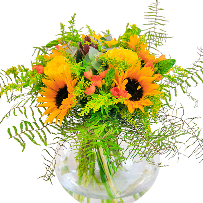 Echoing the rustic grace and golden glow of Cobham's open spaces, this florist-designed bouquet bursts with sunlight and texture. Towering sunflowers with golden petals and rich, coffee-brown centers stand proudly at the heart, radiating warmth and happiness. Gathered close by, sumptuous yellow garden roses spiral open in plush, velvety layers, each bloom a tribute to the cultivated borders along Fairmile. Glossy clusters of red Hypericum berries add bright accents, scattered thoughtfully among lush ferns and trailing green sprigs, evoking the wild beauty of Church Cobham's riverside walks. Subtle yellow filler blossoms and slender, textured spikes are balanced by the muted, silvery tones of eucalyptus, lending freshness and structure. The entire arrangement is showcased in a round, transparent vase, where the interlaced stems create an organic tapestry beneath sparkling water-a symbol of genuine farm-to-vase connection. Designed for moments of celebration or heartfelt congratulations, this bouquet reflects the refined sensibility and down-to-earth spirit of Cobham, available for same-day delivery to homes and events throughout the area.