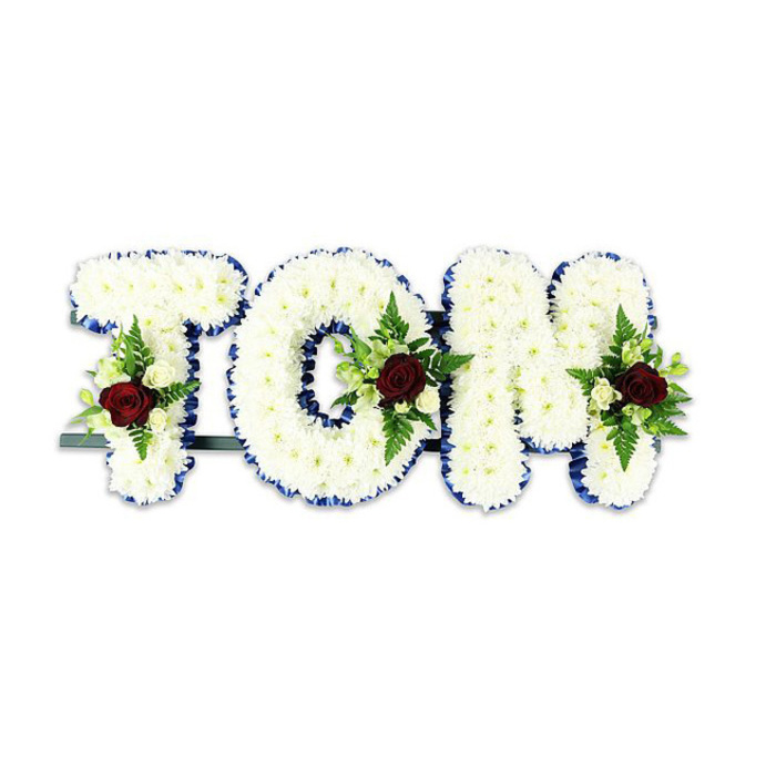 An elegant, formal name tribute fashioned in three large letters that read TOM, each letter densely packed with pristine white chrysanthemums so that the surface becomes plush and tactile. Around every contour a bold royal blue ribbon is tightly applied, forming a crisp, regal edge that separates the letters from the white backdrop and lends a respectful clarity to the message. In the heart or lower area of each character a small, balanced spray sits in quiet prominence: a singular deep red rose in each spray commands attention as the emblem of love and remembrance, while surrounding ivory spray roses and soft green fern foliage cushion the rose and add delicate contrast. The composition is supported by a subtle dark green or black metal bar along the base, visible enough to confirm the arrangement is freestanding and ready to be displayed at a service. Photographed against a clean white surface in soft, neutral light, the arrangement shows fine details - the chrysanthemum petals' gentle ridges, the ribbon's satiny texture, and the glossy sheen of fern leaves - and projects a calm, consoling presence. There is an understated scent profile implied: fresh chrysanthemum coolness layered beneath a faint, warm rose note. As the kind of bespoke floral tribute often commissioned for memorials around Cobham, whether for a service near the River Mole, within walking distance of Cobham Mill, or for a family gathering on the High Street, it speaks of considered practice and care from florists who understand the town's need for dignified remembrance.