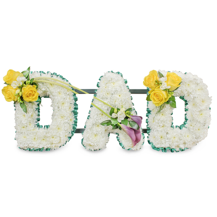 A respectful, handcrafted memorial arrangement spelling DAD in large, freestanding block letters, each letter composed of tightly packed white spray chrysanthemums (or similar small white blossoms) that create a calm, cloud-like surface. The letters are precisely edged with a ruffled dark-green satin ribbon that gives a crisp outline and a subtle reflective sheen against a plain white background. Visible behind the letters are thin dark green metal bars that anchor and align the three letters into a single, stable tribute suitable for chapel display or a graveside service. The left D is highlighted by a cheerful cluster of three to four bright yellow roses, paired with delicate white freesias or gypsophila and sprigs of green foliage; two long, elegant bare green stems extend diagonally from this cluster toward the centre, lending a sense of graceful motion. The centre A carries a more contemplative accent positioned on its lower diagonal: two deep purple or burgundy calla lilies arranged with small white filler blooms and fresh leaves to form a dignified focal point. The right D mirrors the left with an identical yellow-rose cluster on its upper curve. Every selection and placement reflects traditional sympathy symbolism-white for peace and purity, yellow for warm remembrance, and purple for honour-executed with careful wiring and cushioning techniques to maintain shape and freshness. Prepared by a local team familiar with Cobham's funeral customs and frequently arranged for services near Painshill Park, Cobham Mill, or the High Street, this tribute from Flower Delivery Cobham conveys heartfelt respect and lasting remembrance.