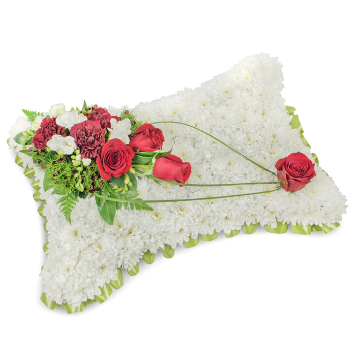 A classic funeral cushion shaped as a gentle rectangular pillow, composed with careful technique so every detail carries meaning. The surface is a dense field of small, immaculate white chrysanthemum blooms; their many layered petals form a velvety, cloud-like plane that evokes peace and repose. Along the slightly raised margins a pale green ribbon and delicate foliage peek through, giving a tidy, understated edge. On the top left corner an opulent cluster of deep red roses and rich burgundy carnations is artfully arranged - the roses in varying stages from tight bud to open bloom, the carnations with their frilled texture - their warm tones creating a strong, respectful contrast to the serene white background. Sprinkled among them are small white freesias or creamy lisianthus, adding soft highlights and a gentle, floral note to the scent profile. Supporting foliage - feathery fern fronds, glossy greens and long bear-grass stems - adds depth and a sculptural sweep: three slender blades of bright green foliage arch across the chrysanthemum surface toward the lower right, guiding the eye in a graceful diagonal. This path is punctuated at its terminus by a single, perfectly formed red rose that appears to rest on the pillow's corner, a quiet emblem of enduring love and remembrance. Photographed in soft, natural light so textures and tones remain true, the cushion reads as an elegant sympathy tribute suitable for services in Cobham and surrounding villages, offering a composed, heartfelt expression of honour and solace.