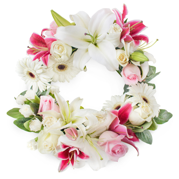 A serene circular wreath of funeral flowers rests against a bright white background, the open ring of blooms creating a gentle halo in tones of white, blush pink, and vivid magenta, much like a composed tribute one might choose for a service in Cobham's village church or a gathering in a local chapel of rest. The design is anchored by large white lilies, each bloom opening widely with soft, slightly ruffled petals and a subtle sheen, their green pistils and golden stamens giving them a refined, almost sacred presence. Interwoven with these are deep pink and fuchsia lilies, their star-shaped petals and occasional speckling adding movement, colour, and a touch of drama, without disturbing the wreath's quiet dignity. White gerbera daisies appear throughout, with layered petals radiating around dark, defined centres, bringing a contemporary feel and a suggestion of hope. Between the larger blooms sit clusters of soft pink roses, some in tightly furled buds and others gently unfolding, offering a tender, personal note of remembrance. Creamy white roses, many fully open with closely packed petals, add volume and a sense of timeless elegance, evoking classic English garden flowers. Smaller white filler flowers, likely miniature carnations or spray-type blooms, fill in every space, creating a plush, continuous ring of petals. The flowers are cradled by lush green foliage, a mix of glossy and matte leaves that wind around the circle, giving depth, structure, and a natural frame reminiscent of Cobham's tree-lined roads and gardens. The open circular shape signifies eternity and unending love, making this wreath a poignant choice for funeral and memorial flower delivery across Cobham, a thoughtful expression of sympathy crafted with care and local expertise.