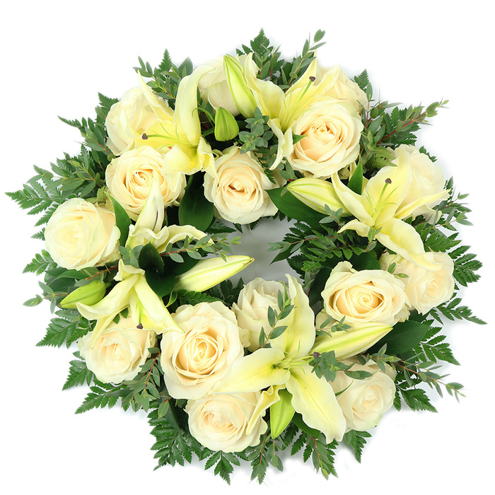 Seen from above and presented on a stark white background, this symmetrical memorial wreath unites creamy white roses and pale yellow lilies with lush green foliage to form a composed and comforting tribute. Approximately fifteen to twenty fully opened roses form a prominent base, their tightly furled centers and layered outer petals giving a sculptural, ivory presence that captures soft daylight. Interspersed are trumpet-shaped lilies in a tender lemon hue; several are fully unfurled, showing graceful, arching petals and delicate light stamens, while others remain in bud, introducing a quiet sense of unfolding and hope. The supporting greenery is intentionally textural: filigree fern fronds lend a soft, feathered outline, while broader, rounded leaves-reminiscent of eucalyptus-provide darker, cooler tones that frame and lift the pale blossoms. The wreath's dense packing produces a lush, almost cushion-like volume, and its perfect circular form communicates lasting remembrance and continuity. The arrangement's tones read cool and calming: muted ivories, soft yellows, and varied greens blending without harsh contrast, making it well suited for a funeral chapel, a graveside at a Cobham cemetery, or as a memorial placed in a quiet corner of a home. A practiced florist's hand is evident in the balance of textures and the restrained scent profile-the roses' gentle sweetness moderated by the lilies' fresh, green edge-creating a composition that expresses respect, solace, and an understated elegance rooted in local understanding of Cobham's gentle landscapes and community grief rituals.