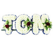 The photograph features a traditional funeral flower arrangement spelling out the name TOM in bold block letters, the sort of personalised tribute often ordered in Cobham for family services and quiet gatherings of remembrance. Each letter is densely covered with immaculate white chrysanthemums, the small, rounded blooms arranged so tightly that they form a uniform, cushion-like surface. The white petals give the design a tranquil, respectful feel, softened further by the subtle play of light and shadow across the textured surface. Framing every letter is a carefully pleated royal blue satin ribbon, its smooth sheen and deep colour providing a clear outline that separates each shape from the plain white background. At considered points along the tribute-on the left of the T, tucked into the curve of the O, and towards the right edge of the M-small sprays of accent flowers break through the white chrysanthemum base. Deep, velvety red roses form the heart of these sprays, their rich hue symbolising enduring love and remembrance. They are supported by tiny white or cream spray roses and slender, starry white blooms reminiscent of freesia, which add grace and lightness. Sprays of fresh greenery, including fern fronds and glossy leaves, fan gently around the roses, contributing depth and a natural movement that recalls the green lanes and woodland edges around Cobham and Oxshott. The letters are aligned on a narrow, dark support, suggesting they are ready to be placed beside the coffin or at the front of a chapel. Overall, the tribute feels dignified and deeply personal, a carefully composed floral memorial created to honour Tom's life and offer comfort to grieving family and friends in Cobham.
