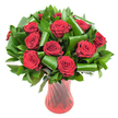 A carefully composed bouquet of twelve to fifteen deep crimson roses sits in the frame, each bloom open to reveal velvety, spiralled petals whose edges catch the soft, even studio light. The roses read as almost wine-dark at their core, graduating to luminous red at the petal tips; they are clustered into a lush, rounded dome that feels both abundant and intimate. Nestled among them is a generous mass of dark green foliage - glossy salal or ruscus leaves form a dense, matte backdrop while broader aspidistra-style leaves have been folded, rolled and curved into elegant cylindrical loops that arc around the stems like quiet architectural gestures. Those sculpted green elements add surprising modernity and structural rhythm to a classic rose presentation, creating shadowed channels that deepen the colour contrast. The bouquet is anchored in a tall, translucent ruby-red glass vase whose subtle ribbed or twisted texture throws vertical highlights and reflections, echoing the roses' passionate tones and lending a refined, tactile counterpoint to the soft petals. Everything is set against a clean white background so the reds and greens sing clearly, the lighting emphasising fine petal veins and the sheen of each leaf. A faint, sweet rose scent seems to rise from the tight cluster, suggesting a presence beyond the image - an anniversary gift or a romantic centrepiece intended to fill a small Cobham drawing room or a table at a celebration in nearby Painshill Park. The overall effect is one of considered luxury and warm intimacy: traditional romance rendered with a contemporary, local florist's restraint and attention to form and texture.