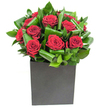A compact, modern composition of deep crimson roses set against a backdrop of glossy, sculpted greenery, photographed in bright, neutral light that highlights the velvety texture of each bloom. The roses open in generous spirals, their petals layered like satin, while broad emerald leaves have been carefully curled and overlapped to form a rhythmic frame that both supports and accentuates the flowers' intense colour. The bouquet sits in a matte black, square box vase whose straight edges and low profile give the arrangement a grounded, contemporary silhouette - the contrast between the soft blooms and the vessel's clean geometry feels deliberate and poised. The lighting is crisp and even, catching subtle highlights on the leaf surfaces and lending the red tones a rich, almost tactile glow, as if the roses had been unfurled moments earlier. Close to the arrangement you might imagine a faint perfume of classic rose softened by green, almost citrus-like notes from the foliage; the scent reads as fresh rather than heavy. As a local florist I see this piece fitting equally well on a low dining table in a Cobham home or as a thoughtful gift collected from Cobham High Street - it reads as both romantic and quietly modern. The design's balance of structure and lushness makes it suitable for anniversaries, heartfelt thank-yous, or to give an everyday room a moment of refined drama. Every crease in the petals and every curl of leaf reflects deliberate craftsmanship, an unspoken promise that the flowers were handled with care from stem to vase.